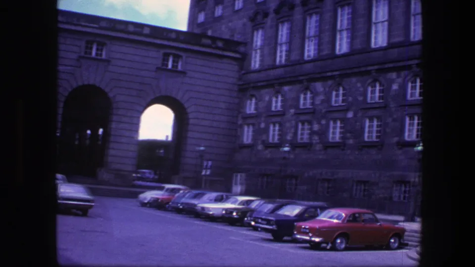 1975 Copenhagen view of a large arched gateway, adjoining building facades, and rows of parked cars including a red car in the foreground.