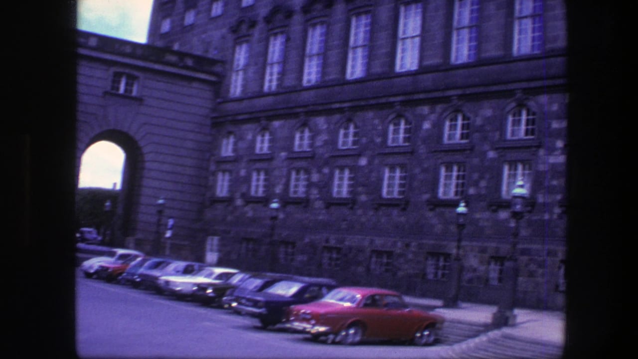1975 Copenhagen view of a large stone building, an arched passage, and a row of parked period cars.