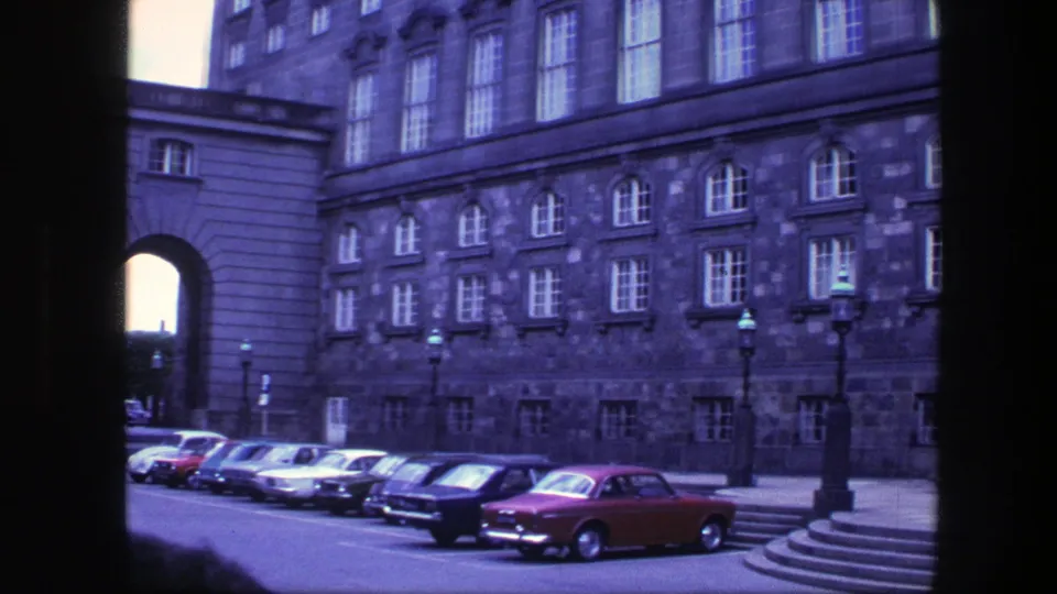 1975 Copenhagen street view with multiple parked period cars, a red car, and a large stone building behind them.