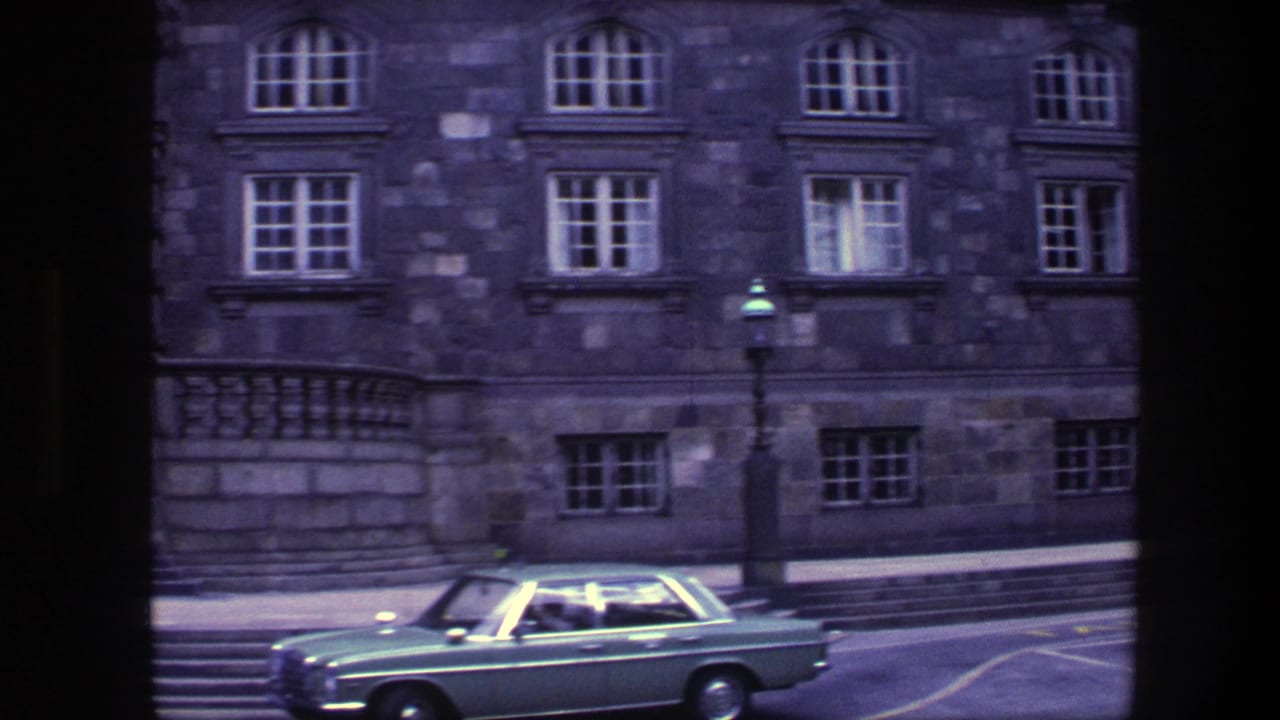 Green sedan passing a large stone building with rows of windows and streetlamps.