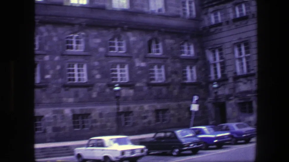 A white car and several parked vehicles sit before a large Copenhagen stone building, with a small pedestrian visible near the facade.