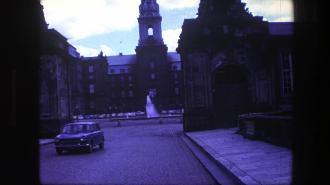 1975 Copenhagen 8mm frame showing a small car on a roadway between historic stone gateways, with a fountain and tower beyond.