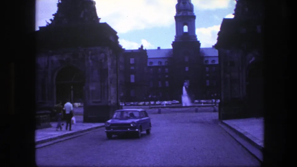 1975 Copenhagen 8mm frame with a small car, two distant pedestrians, stone gateway, fountain, and tower in a city setting.