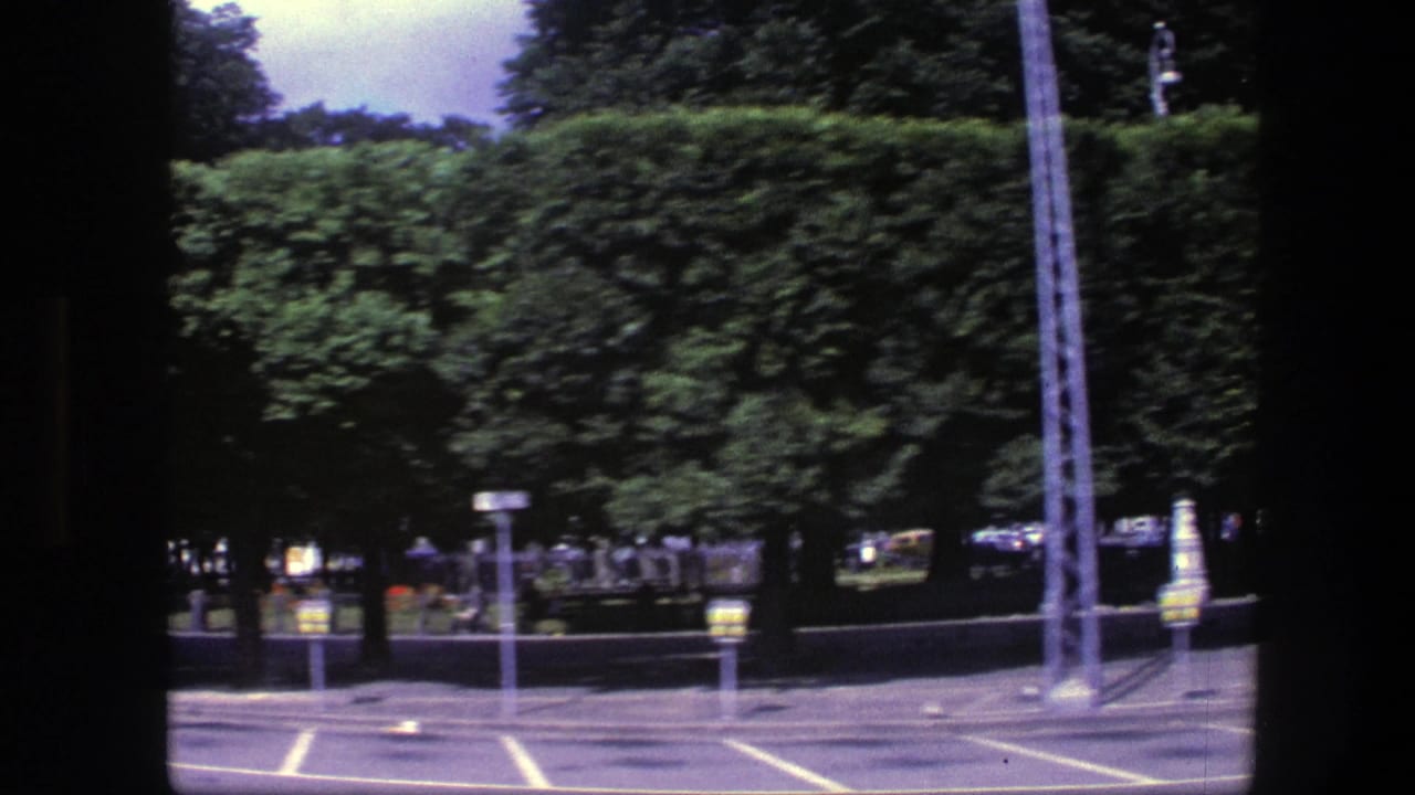 1975 Copenhagen home-movie frame showing marked parking spaces near a long green hedge, trees, and street fixtures.