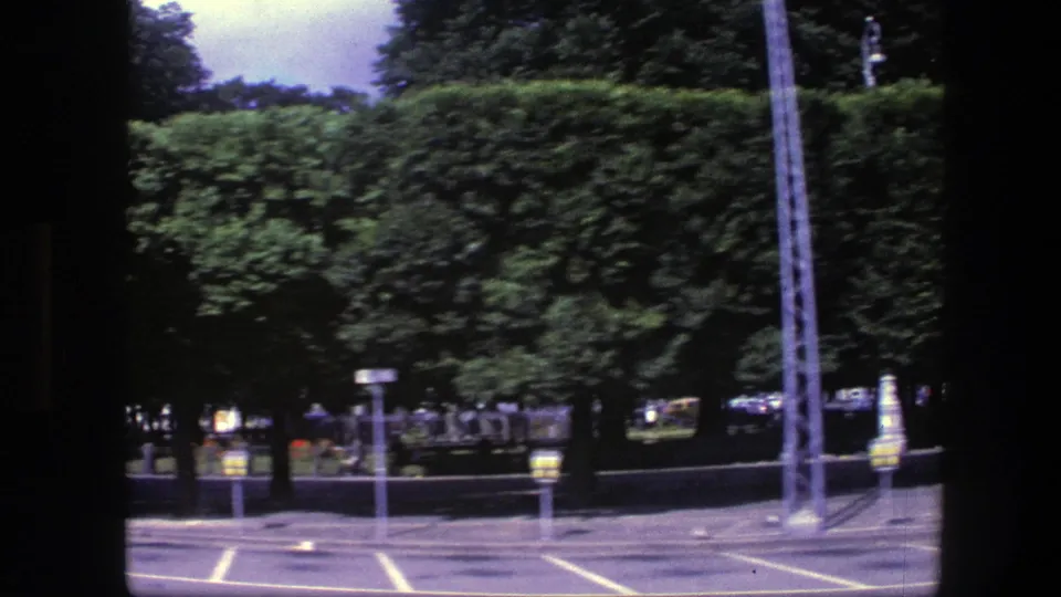 1975 Copenhagen home-movie frame showing marked parking spaces near a long green hedge, trees, and street fixtures.