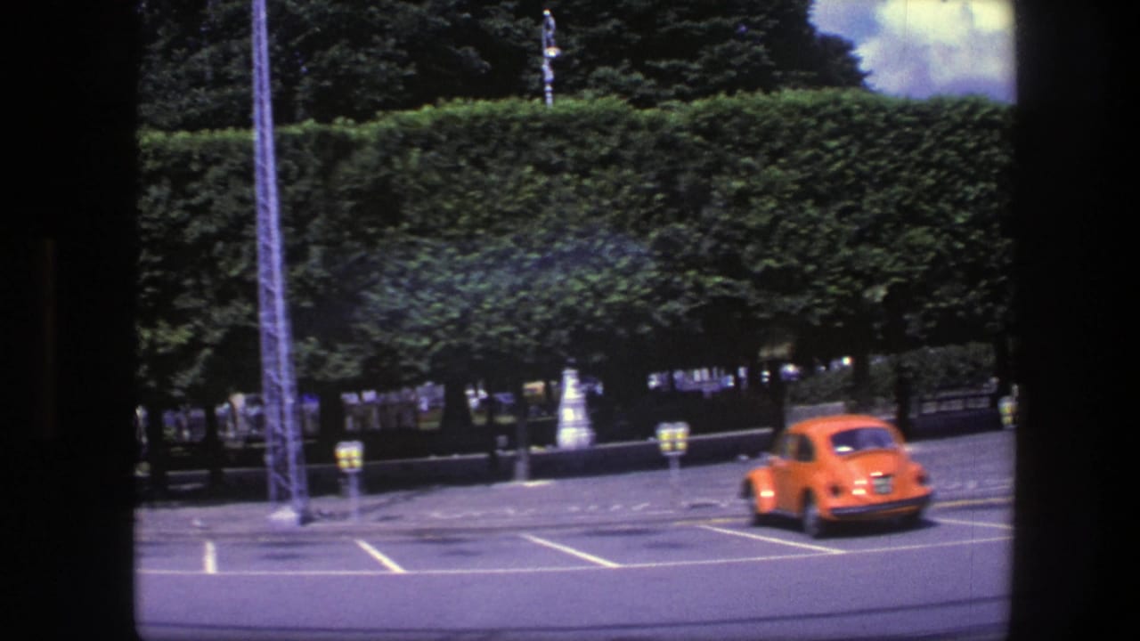 Orange compact car drives through a parking area beside a long green hedge.