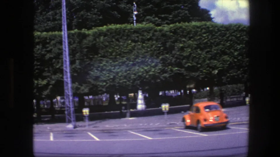1975 Copenhagen frame with an orange compact car moving across a marked parking area near clipped trees and a hedge.