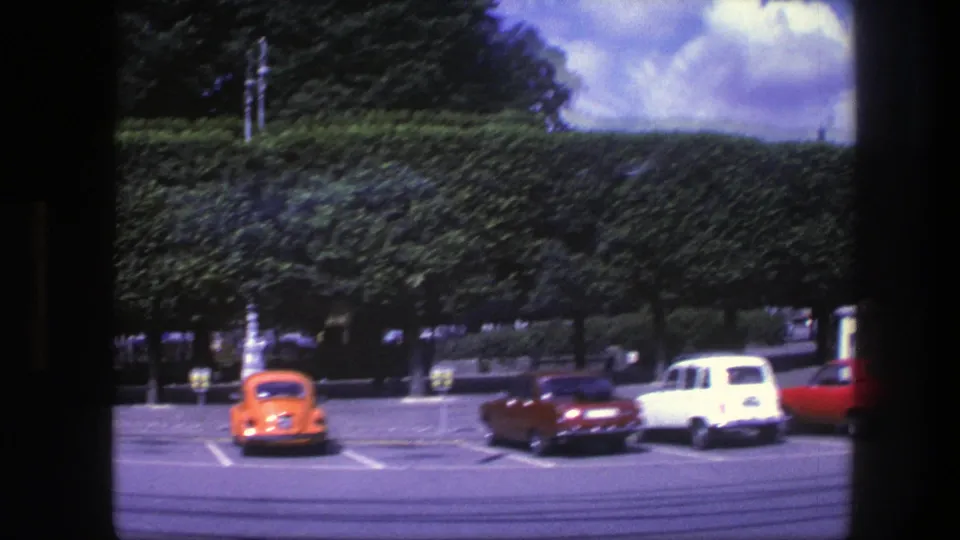 1975 Copenhagen home-movie frame showing an orange car, red car, white wagon, and another red car parked near a clipped hedge.