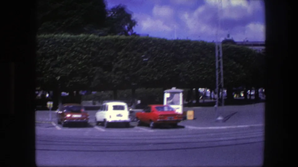 1975 Copenhagen frame showing red and white parked cars, a trimmed hedge, street lines, and a metal mast in the distance.