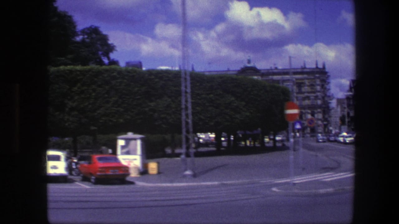 Street corner with clipped hedges, no-entry signs, a red car, and gray buildings beyond.