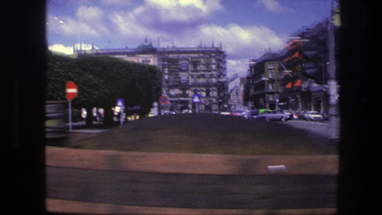 Moving 1975 Copenhagen street view past clipped hedges and no-entry signs toward ornate multi-story buildings with light traffic in the distance.