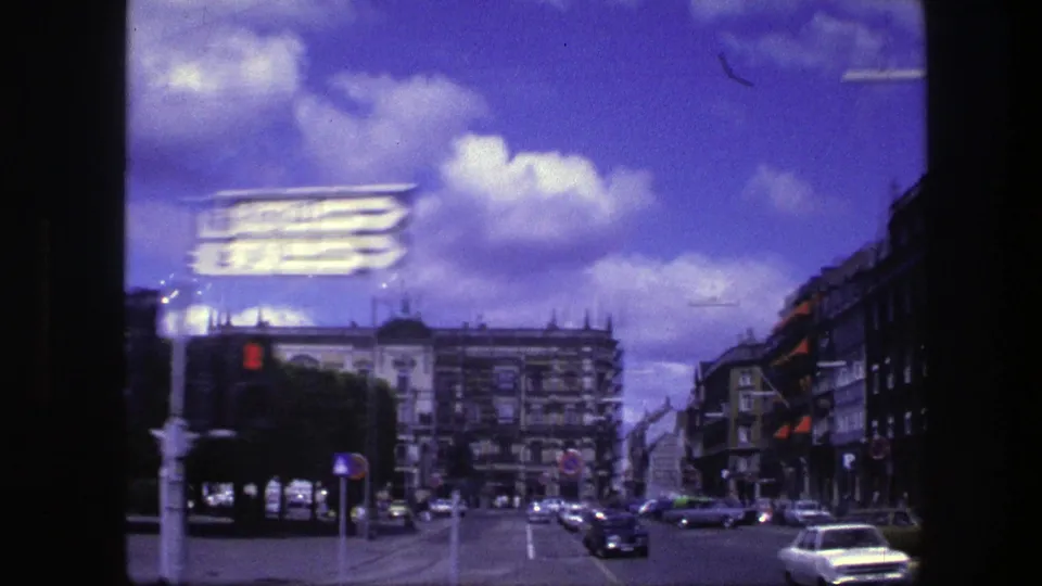 1975 Copenhagen street scene with gray building facades, traffic signs, moving cars, and a bright sky with scattered clouds.