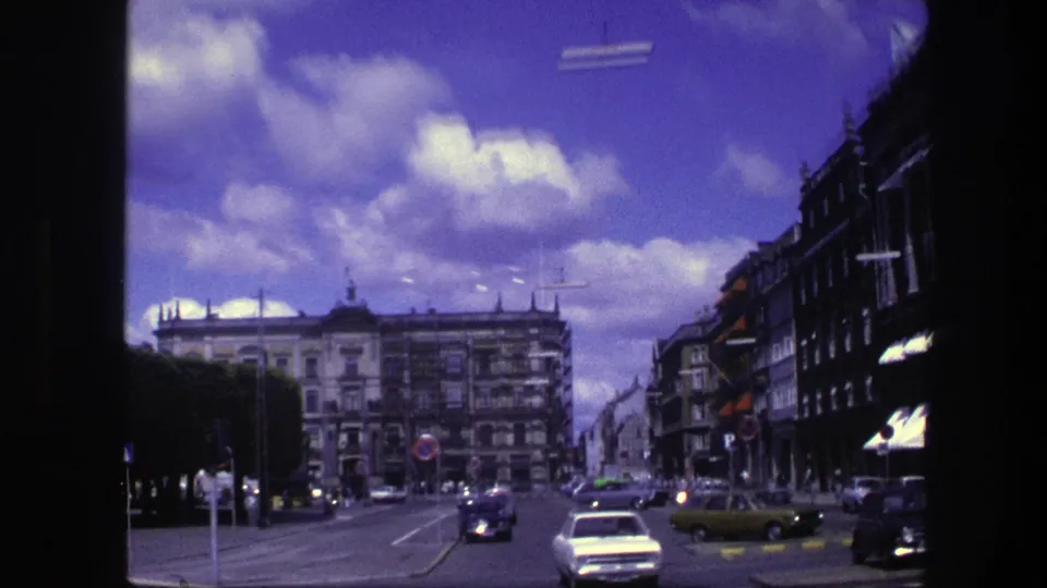 1975 Copenhagen street view with a broad roadway, light car traffic, clipped hedges, formal building facades, and bright clouds above.