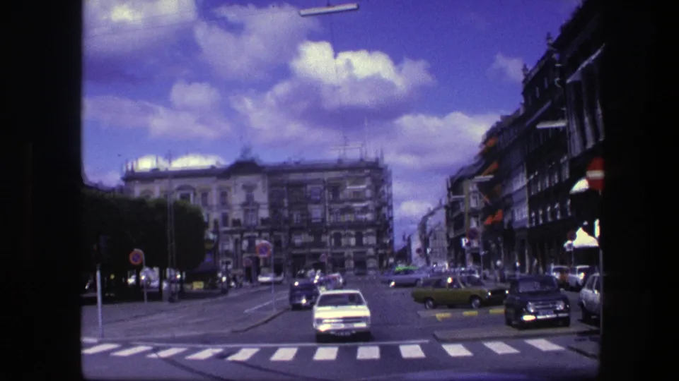 1975 Copenhagen street scene with classic cars crossing a wide intersection beneath a bright sky and rows of urban buildings.