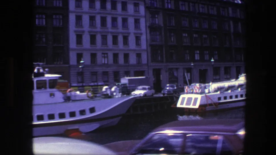 Copenhagen harbor frame with a ferry bow, parked cars along the quay, tall buildings, and a dark passing car at the bottom edge.