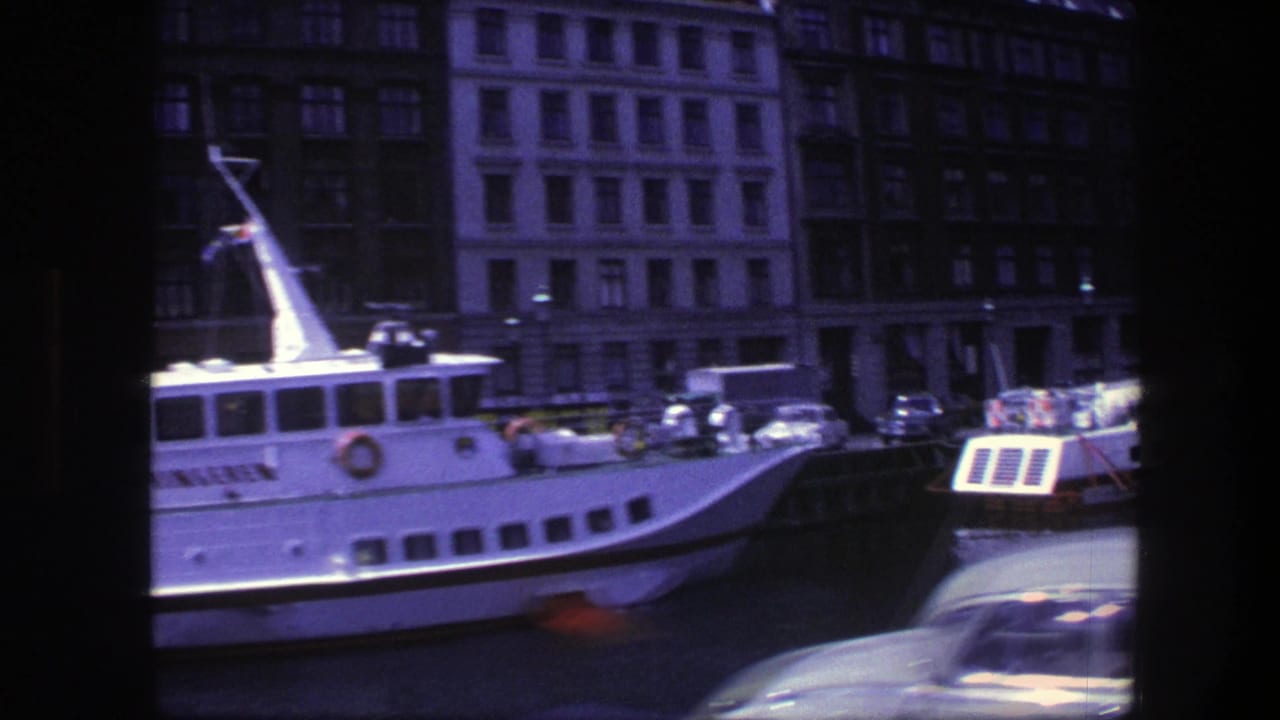 White passenger boat docked at a Copenhagen harbor beside classic cars and tall waterfront buildings.
