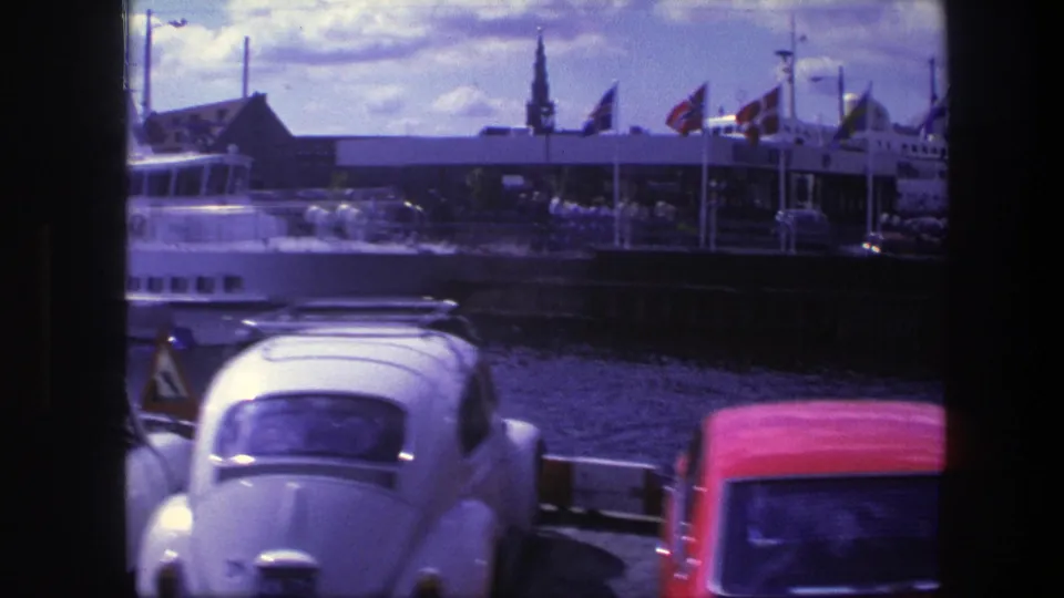 Copenhagen harbor in 1975 with the passenger boat partly passing frame, flags on the quay, and classic cars including a white Beetle and red sedan.