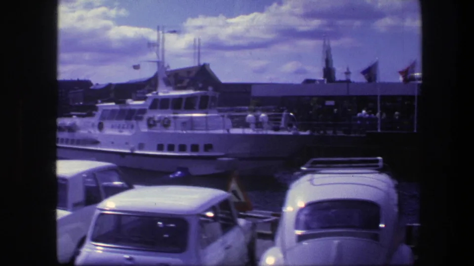 1975 Copenhagen harbor view with parked cars, a white passenger boat, terminal buildings, flags, and cloudy sky seen through 8mm frame edges.