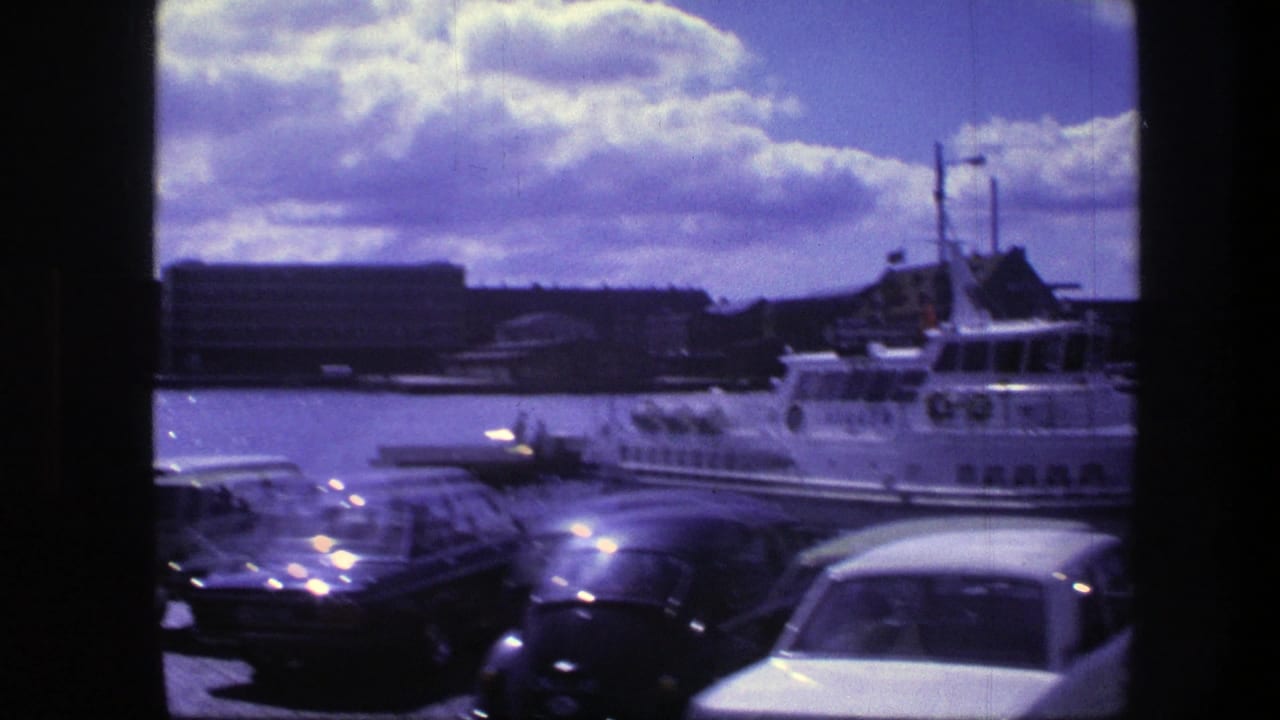 1975 Copenhagen waterfront scene with parked cars in the foreground and a white passenger boat near harbor buildings under a cloudy sky.