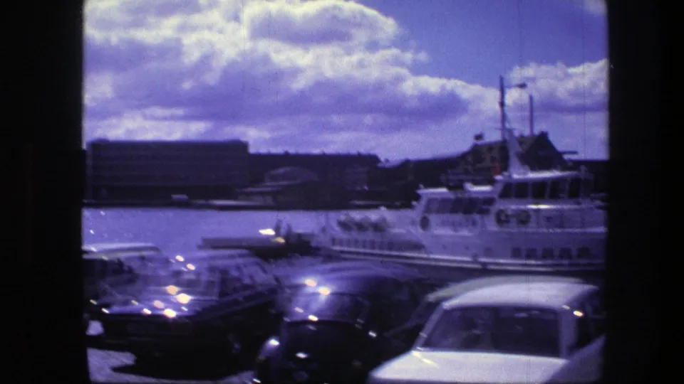 1975 Copenhagen waterfront scene with parked cars in the foreground and a white passenger boat near harbor buildings under a cloudy sky.