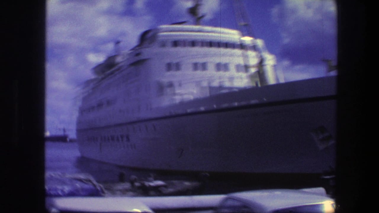 1975 Copenhagen harbor view of a large white passenger ship, with classic cars low in the foreground and cloudy sky above.