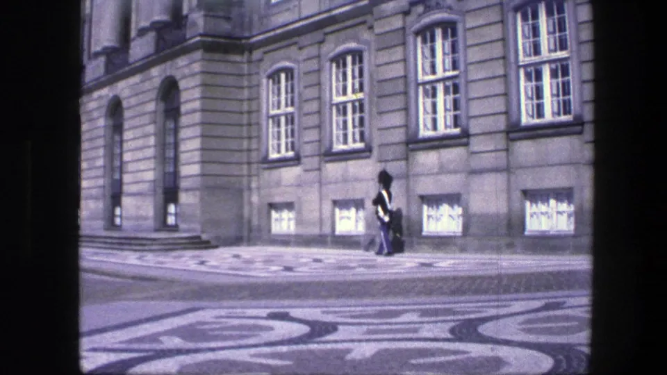 Copenhagen street view with a uniformed guard walking past a formal stone facade, arched windows, and patterned paving.