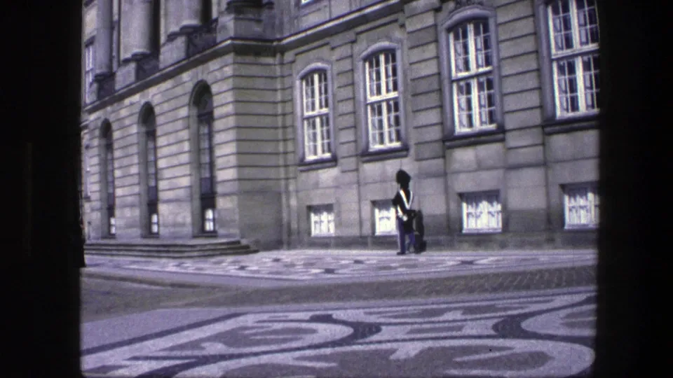 Final Copenhagen frame showing a uniformed guard by a formal stone building, with tall windows and decorative paving visible.