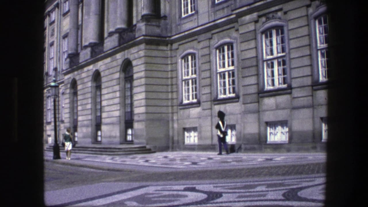 Guard stands near a stone building while a civilian walks in the background.
