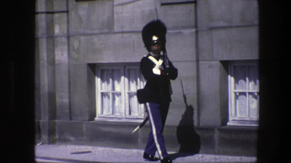 Closer 8mm frame from Copenhagen shows the royal guard in dark uniform and white cross straps standing near curtained basement windows.