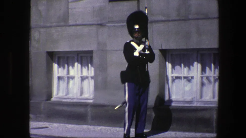 The final 1975 Copenhagen frame shows the royal guard facing forward with rifle, framed by grey stonework, low curtained windows, and strong sunlight.