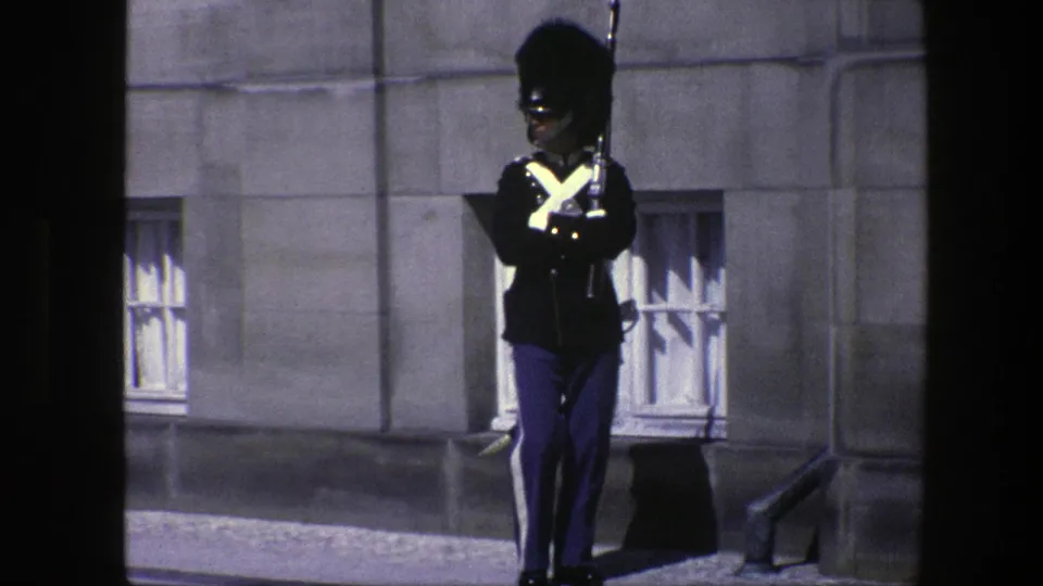 A ceremonial guard in dark uniform and blue trousers stands outside a Copenhagen stone building in 1975.