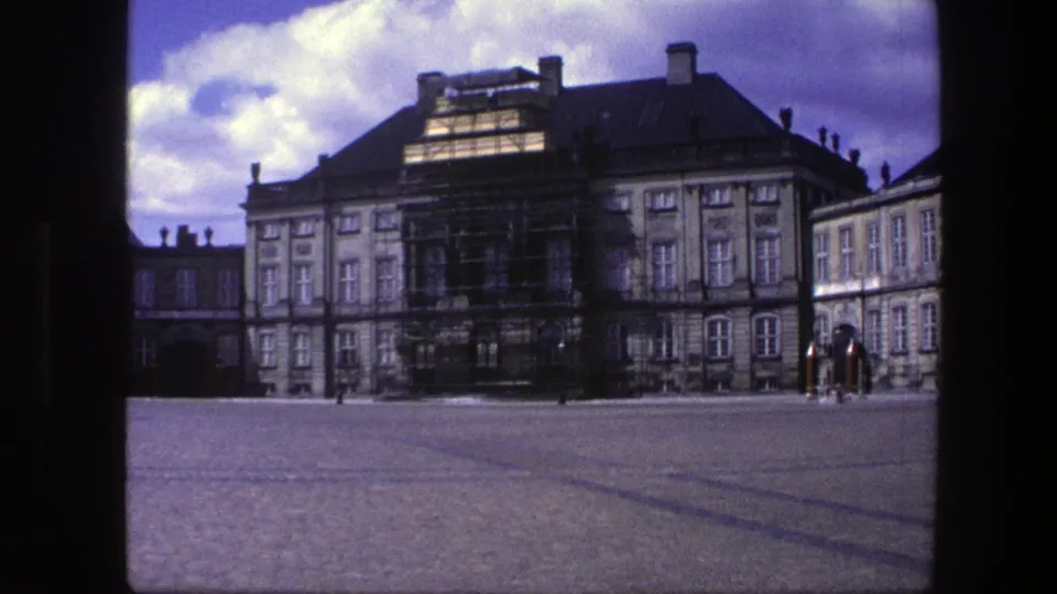 1975 Copenhagen home-movie frame showing a grand palace facade, central scaffolding, cobblestones, and small figures near the entrance.