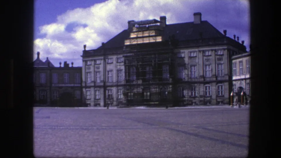 1975 Copenhagen frame showing a palace courtyard, scaffolded facade, cloudy sky, and distant figures near a guard post.