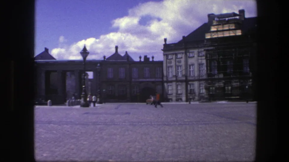 1975 Copenhagen frame showing a wide public square with historic buildings, restoration scaffolding, a streetlamp, pedestrians, and a red car crossing.