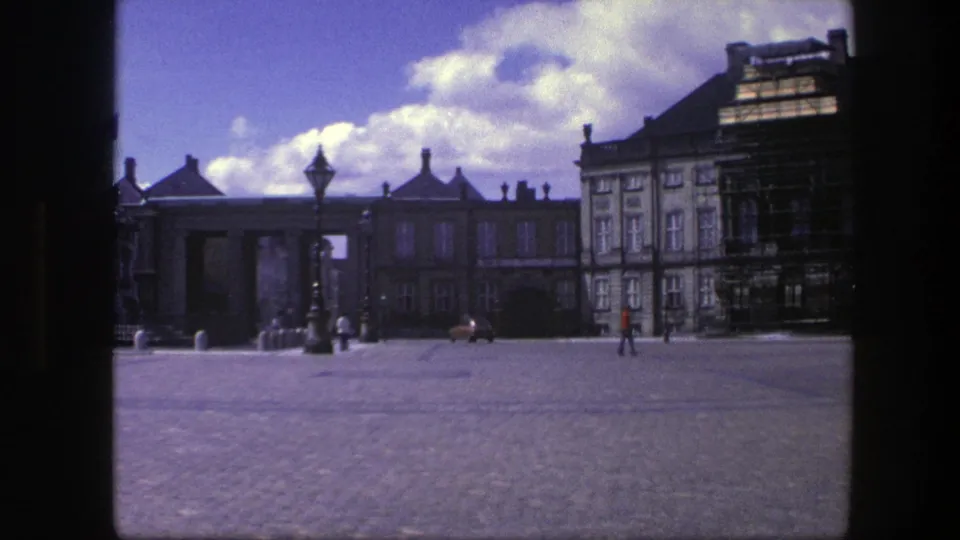 1975 Copenhagen 8mm frame of a broad square with palace architecture, a scaffolded building section, small pedestrians, and a car near the edge.