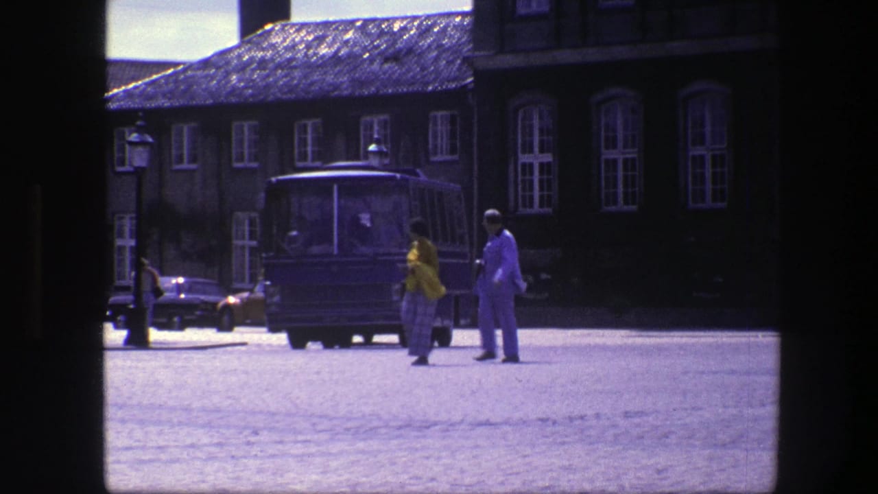 In 1975 Copenhagen, two adults walk across a cobblestone square with a blue bus, cars, lampposts, and dark building facades behind them.