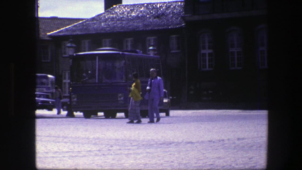 A couple walks near a blue bus on a cobblestone square with older cars behind them.