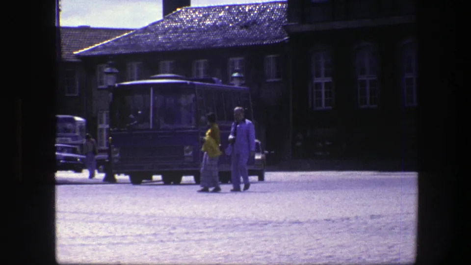 Two adults in bright clothing walk across a Copenhagen cobblestone square in 1975, with a blue bus, older cars, and tall windows in the background.