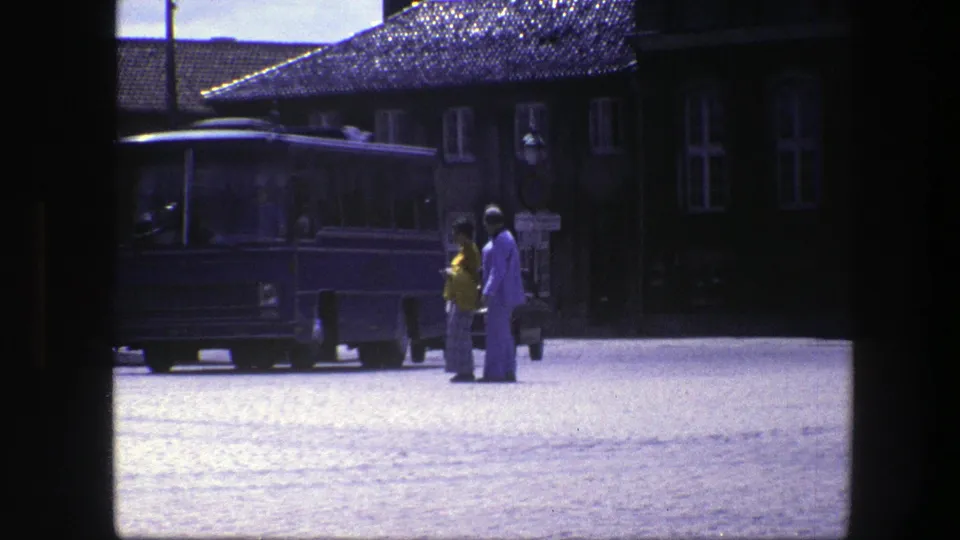 The couple appears closer to the blue bus in a 1975 Copenhagen street scene, framed by cobblestones, building facades, and strong sunlight.