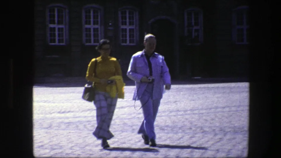 In a 1975 Copenhagen home-movie frame, a woman in a yellow top and plaid trousers walks beside a man in a light suit across cobblestones.