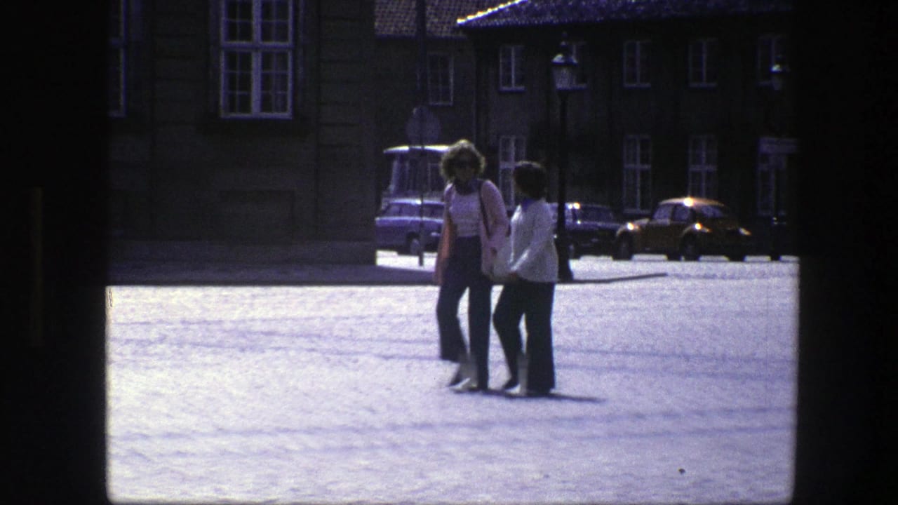 Two adults walking across a paved city square near parked cars and older buildings.