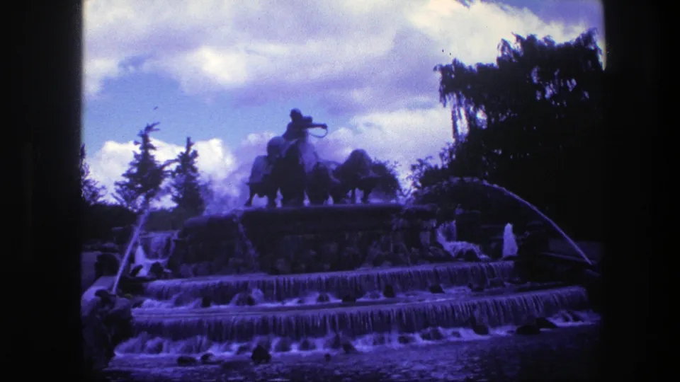 A Copenhagen public fountain appears with stepped cascades, arcing water, trees, clouds, and a silhouetted equestrian statue above.