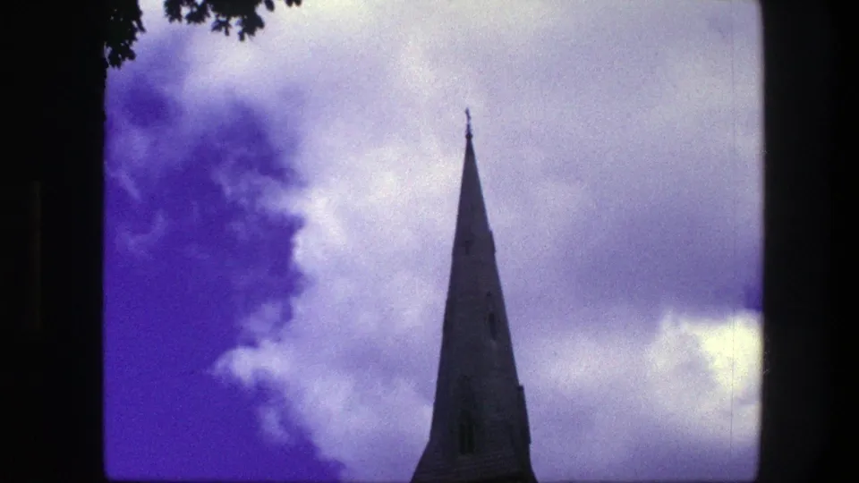 1975 Copenhagen home-movie frame showing a tall pointed spire against dramatic clouds, with tree foliage at the upper edge.