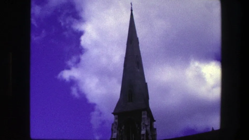 1975 Copenhagen 8mm frame centering a pointed church steeple beneath purple-blue clouds, with soft grain and frame line visible.