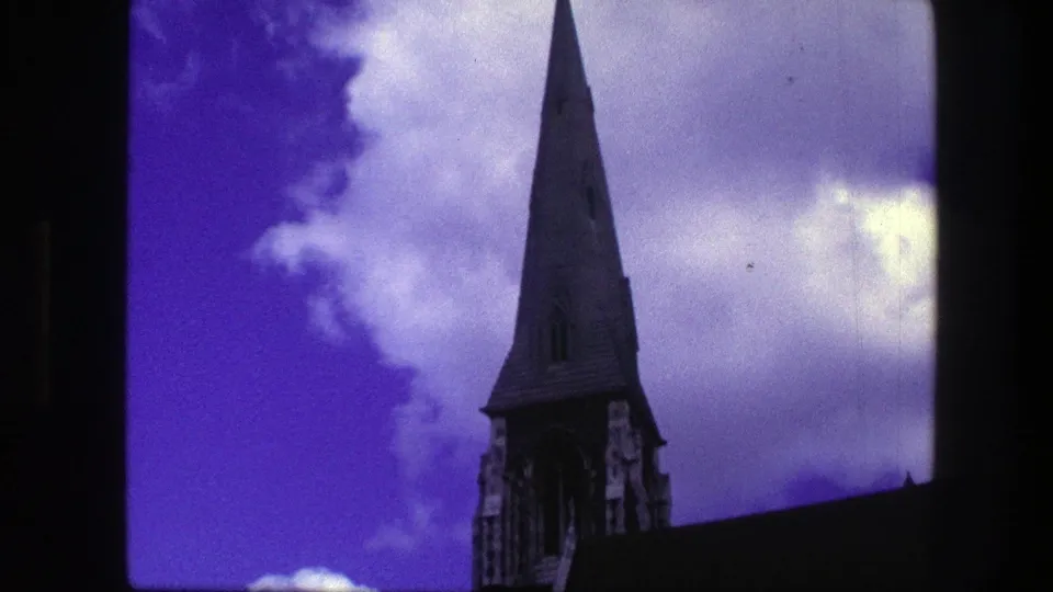 1975 Copenhagen home-movie frame showing the church tower and pointed spire above a dark roofline with purple-blue clouds behind it.