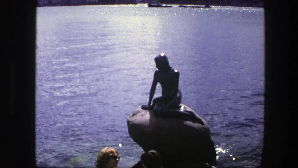 Copenhagen 1975 frame of the Little Mermaid statue seen from a visitor viewpoint, with bright harbor water and partial people at the bottom.