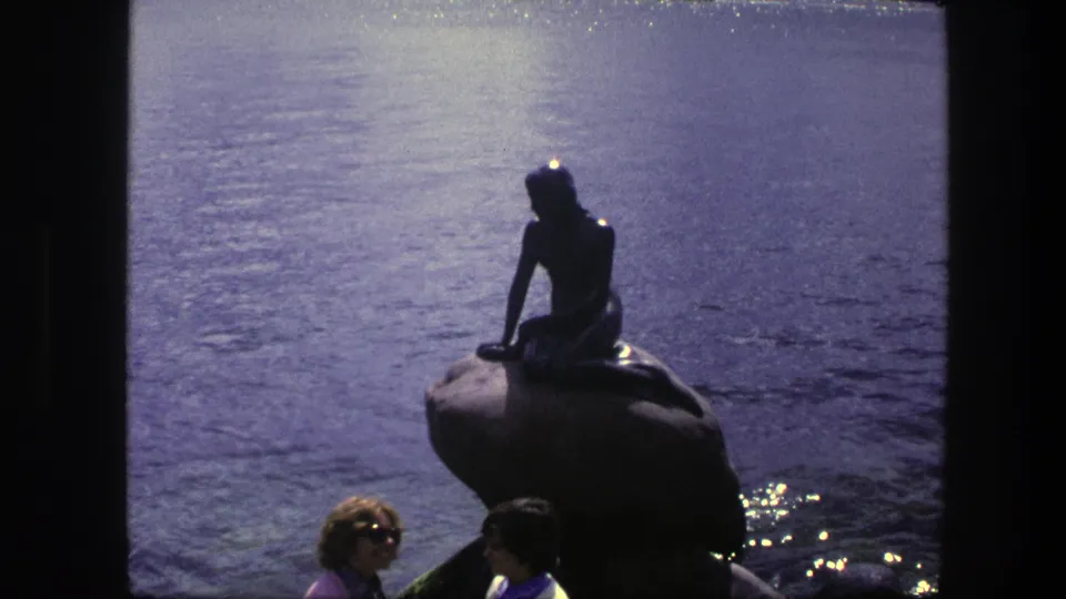1975 Copenhagen 8mm frame showing the Little Mermaid statue on its rock base, with visitors close to the shoreline and sparkling water behind.