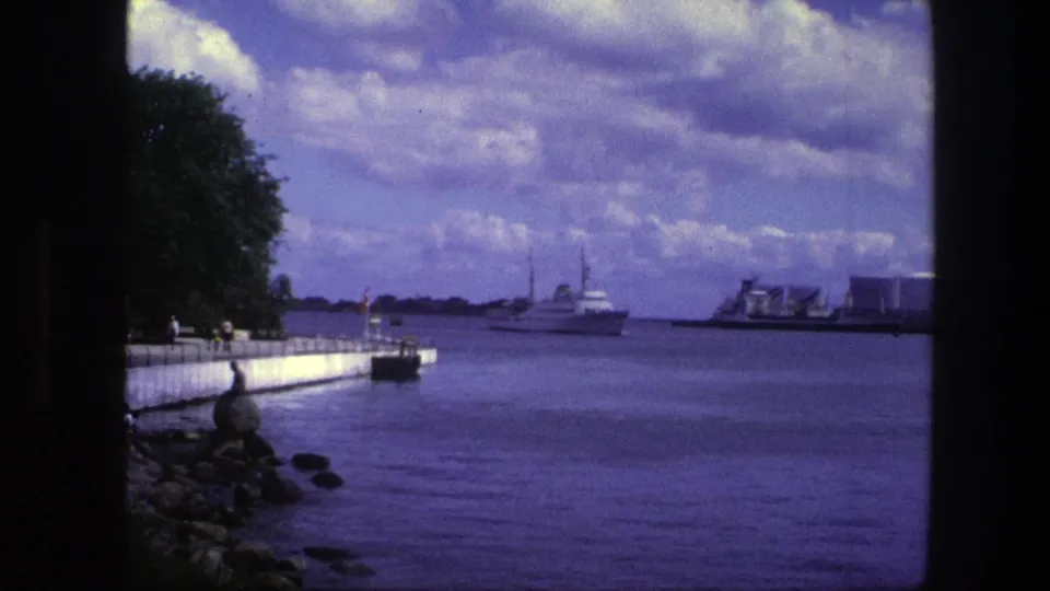 1975 Copenhagen harbor frame showing a waterfront path and trees on the left, with a large ship and smaller vessel on the water under clouds.