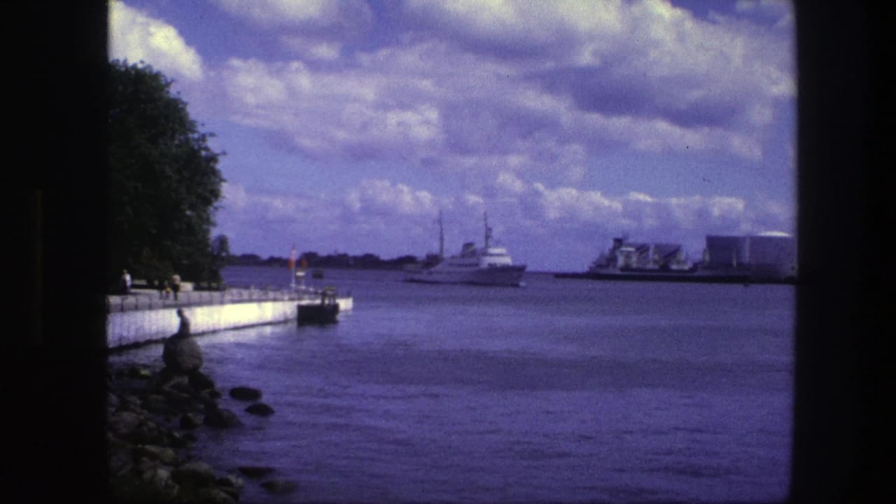 White passenger boat crossing a wide waterway under cloudy sky beside a concrete quay.