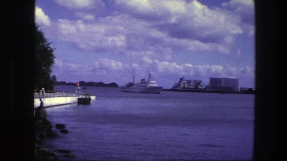 1975 Copenhagen waterfront frame with a white boat, broad water surface, pier edge, rocks, trees, and distant harbor structures.